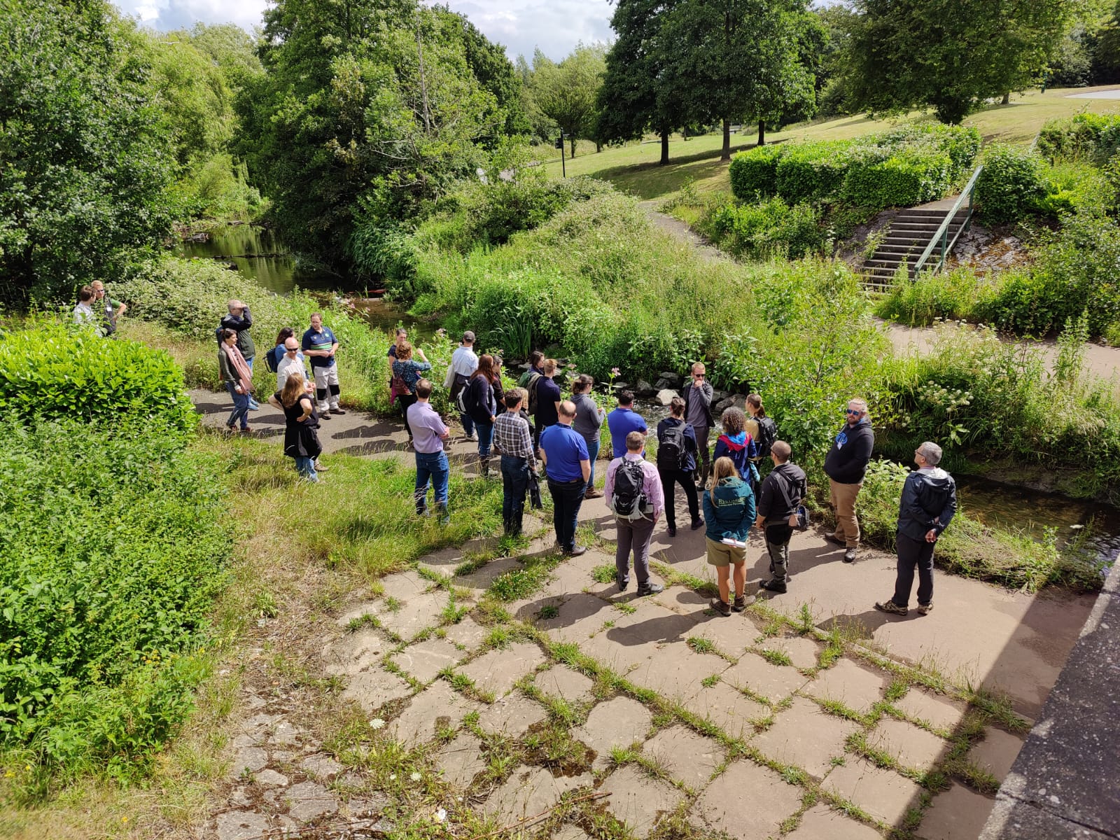 A large group of people at the conference. They are standing next to a river and watching the presenter speak